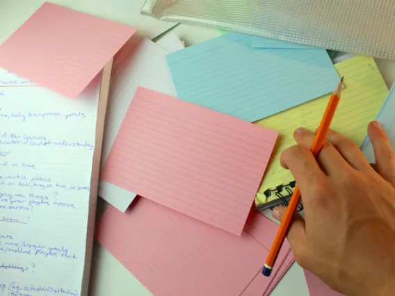 Image of a desk with a notebook and flashcards.  Hand at bottom right with a pencil between thumb and middle finger.