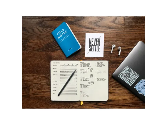 Top view of a desk with an open notebook, half a laptop that has stickers, airbuds, and a sticker that says never settle.