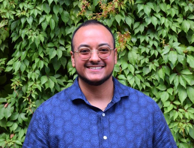 Key Sanchez wearing glasses and a blue shirt in front of a wall with green leaves
